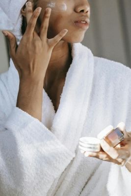 Crop unrecognizable  young female in white bathrobe applying moisturizing cream on face after shower