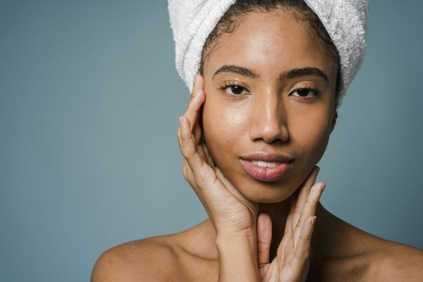 Calm African American female with towel turban on head and bare shoulders touching face and looking at camera against blue background in studio