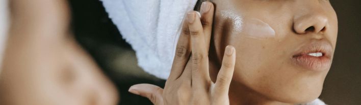 Crop unrecognizable young ethnic female in terry robe applying moisturizing cream on cheek while reflecting in mirror